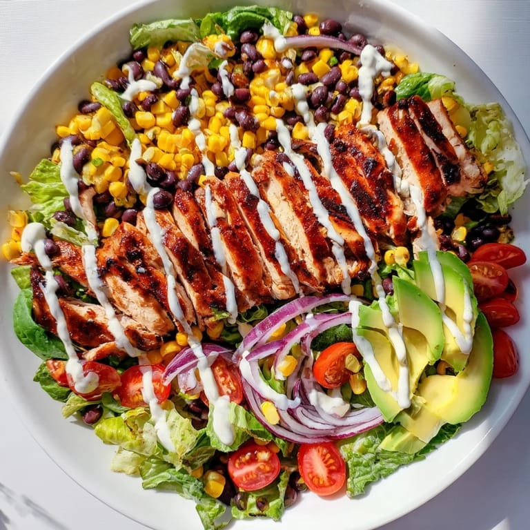 A close-up of BBQ Chicken Salad shows creamy avocado slices and shredded cheese atop the greens, with ranch drizzle glinting in the light.